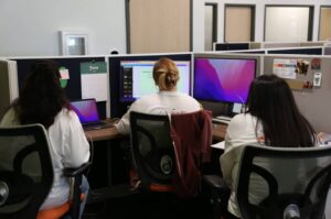 Three women, all wearing the same white shirt) face away, looking at a computer screen making a presentation. 
