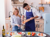 A man and woman cooking together, with the man wearing an apron featuring the "Our Kitchenware" logo